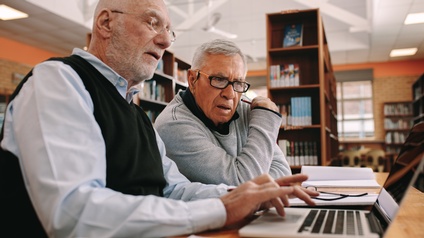Zwei ältere Personen sitzen in einer Bibliothek am Tisch und blicken auf einen Laptopbildschirm, im Hintergrund stehen Bücher in Bücherregalen.