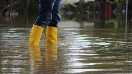 Person in Jeans steht mit gelben Gummistiefeln in tiefem Wasser, Hochwasser