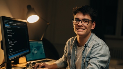 Seitliche Aufnahme einer Person, am Schreibtisch sitzend und auf einer Tastatur tippend. Auf dem Tisch sind ein Bildschirm und ein Laptop. Auf dem Bildschirm ist ein Quellcode. Die Person blickt direkt in die Kamera und trägt eine Brille