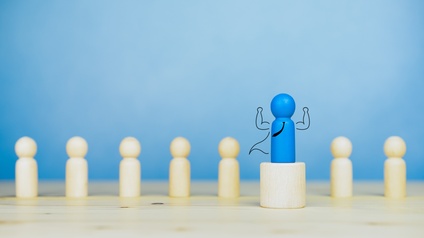 Kleine blaue Holzfigur steht auf Holzpodest, illustriert Krawatte, Umhang und erhobene starke Arme, im Hintergrund weitere naturfarbene Holzfiguren