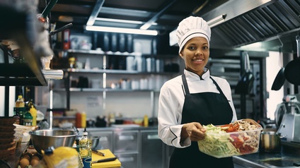 Eine Person mit Kochmütze, Kochkleidung und Schürze steht in einer Gastroküche und blickt freudig in die Kamera. In den Händen hält sie eine Plastikbox, befüllt mit geschnittener Zucchini, Paprika und Pilzen.