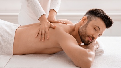 Person mit kurzen braunen Haaren und Bart liegt mit geschlossenen Augen auf einer Liege und genießt eine wohltuende Massage, Seitenansicht