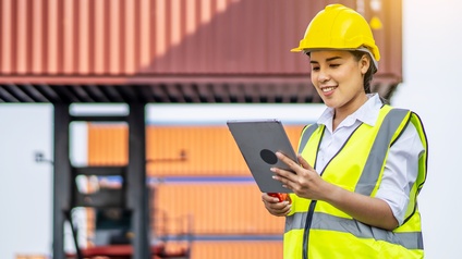 Eine Person in Arbeitskleidung mit helm steht vor Frachtcontainern und hält ein Tablet in der Hand, auf das sie lächelnd blickt.