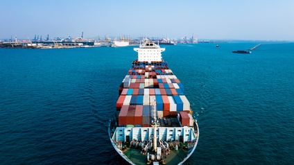 Ein Frachtschiff mit vielen Containern, das auf einem Gewässer fährt. Im Hintergrund ist ein Hafen mit vielen Hafenkränen