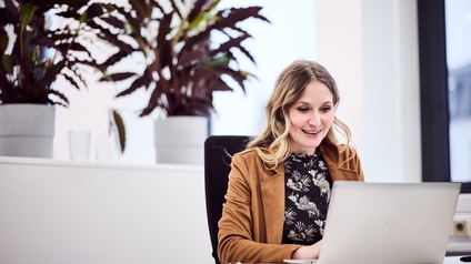 Eine Person sitzt vor ihrem Laptop und tippt in die Tastatur. Hinter ihr ist ein Regal, auf dem Topfpflanzen stehen.