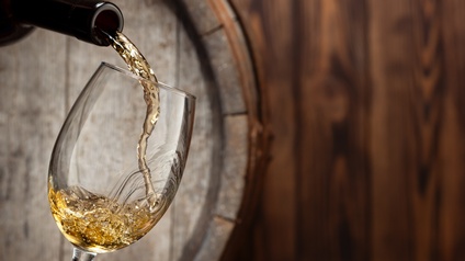 In der linken Bildhälfte wird aus einer Flasche Weißwein in ein Weinglas geschenkt. Hinter Flasche und Glas ist ein liegendes Holzfass