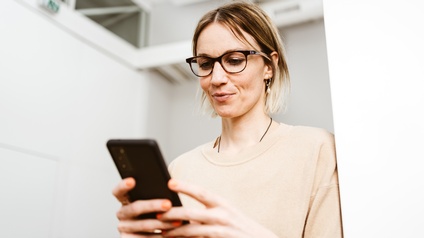 Nahaufnahme einer Person mit Brille, die lächelt, sodass sich Grübchen neben dem Mund bilden. Ihr Blick ist auf ein Smartphone gerichtet, das sie in beiden Händen hält. Sie steht in einem sterilen Raum