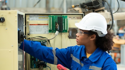 Person mit Schutzbrillen, weißem Helm und blauem Arbeitsoverall in Seitenansicht kontrolliert Elektrokasten, im Hintergrund Türe des Kasten mit Kabelkanälen und Kabeln