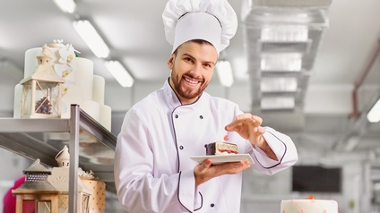 Person in weißer Arbeitsbekleidung steht in einer Backstube und hält ein Gebäck in der Hand