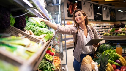 Lächelnde Person in Lebensmittelgeschäft greift in Gemüseregal nach Häupel Salat, hält in einer Hand Tablet, vor ihr voller Einkaufswagen