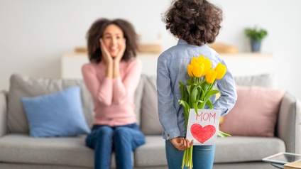 Kind steht vor einer erwachsenen Person, die auf einer Couch sitzt und hält einen Strauß gelber Tulpen sowie eine Geschenkkarte mit der Aufschrift MOM und einem gezeichnetem Herz