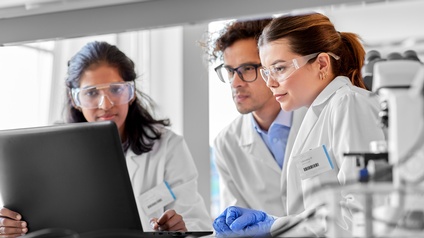 Drei Personen mit Brillen in weißen Kitteln blicken auf aufgeklapptes Notebook, im Vordergrund verschwommen Mikroskop