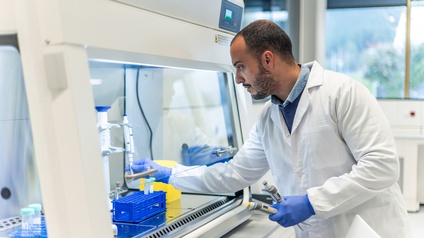 Seitliche Aufnahme einer Person mit weißem Kittel, Bart, dunklen Haaren und Handschuhen. Die Person steht an einer Station mit einer Glasscheibe, hinter der Reagenzgläser stehen