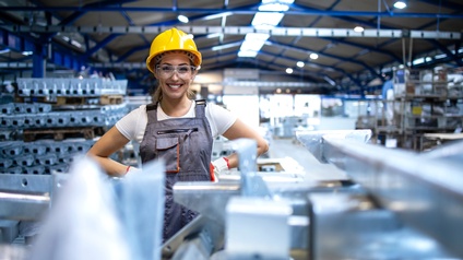 Lächelnde Person in Arbeitskleidung mit gelben Schutzhelm und Schutzbrille steht in einer industriellen Produktionsstätte