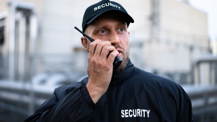 Frontale Aufnahme einer Person mit Bart, dunkler Jacke und dunkler Kappe, auf denen jeweils Security steht. Die Person hält sich ein Walkie Talkie vor den Mund. Im Hintergrund ist ein Gebäude