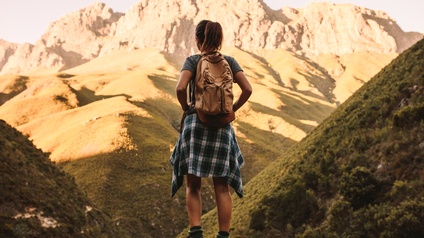 Person mit Rucksack und in Wanderkleidung steht in Rückenansicht auf Fels vor Berg- und Wiesenpanorama
