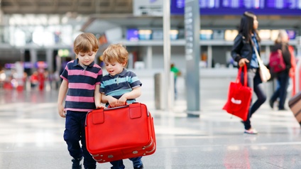 Zwei Kleinkinder mit gestreiften Shirts und dunklen Hosen gehen eine Halle entlang, einer von beiden trägt einen roten Reisekoffer, im Hintergrund befinden sich weitere Personen mit Gepäck sowie eine große blaue Anzeige