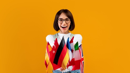 Lächelnde Person steht vor einer gelben Wand. In den Händen hält sie verschiedene Länderflaggen, zum Beispiel Spanien, Türkei, Deutschland, Kanada