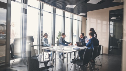 Gruppe an Personen in Businesskleidung befindet sich rund um einen Besprechungstisch in einem modernen und hellen Besprechungsraum