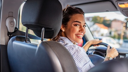 Lächelnde Person mit langen dunklen Haaren und gestreifter Bluse sitzt in einem Auto und blickt auf die Rückbank zurück