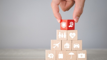 Holzwürfel stehen zu einer Pyramide gestapelt auf einer hölzernen Oberfläche. Auf den Würfeln sind Symbole, zum Beispiel ein Haus, ein Auto und ein Regenschirm. Der oberste Würfel ist rot und wird vom Zeigefinger und Daumen einer Person gehalten