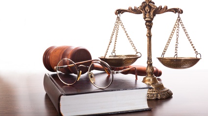 Auf einem braunen Holztisch sind Objekte platziert: Eine goldene Waage mit Verzeihrungen, ein Buch mit braunem Einband, eine Brille und ein Hammer. Der Hintergrund ist weiß.