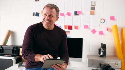 Lächelnde Person sitzt auf Schreibtisch und bedient Tablet, im Hintergrund Fotografieutensilien und Post-its an der Wand