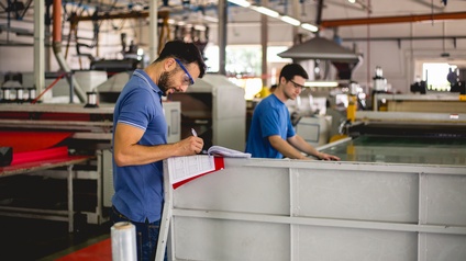 Zwei Personen in großer Industriehalle an Produktionsmaschine für Kunststoff stehend, einer Person im Fokus nimmt Notizen vor