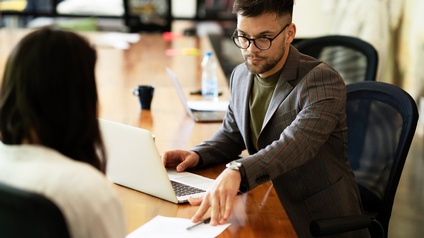 An einem Tisch sitzen zwei Personen. Die rechte Person schiebt der anderen Person einen Zettel und einen Stift hin
