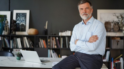 Halbporträt einer auf einem Tisch sitzenden Person in einem hellblauen Hemd und dunkler Hose, die vor dem Oberkörper die Arme verschränkt hat. Auf dem Tisch ist ein aufgeklappter Laptop. Im Hintergrund ist ein niedriges Regal mit Büchern