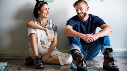 Zwei Personen sitzen nebeneinander auf Plastikfolie, die auf einem Holzboden ausgelegt ist. Die Personen lehnen sich an der Wand hinter ihnen ab und lächeln. Auf der Wand sind verschiedene Farbstriche. Die linke Person trägt eine Latzhose mit Farbklecksen