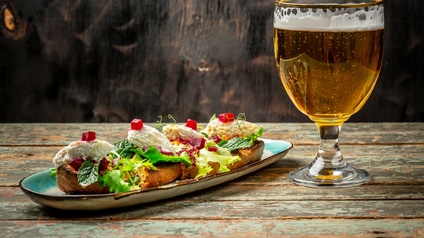 Auf einem Holztisch steht ein Glas befüllt mit Bier. Links daneben steht ein Teller mit vier belegten Brötchen