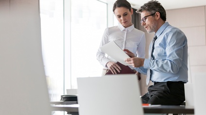 Zwei Personen in Businesskleidung besprechen Unterlagen in einem Büro, eine Person trägt eine Brille sowie einen Bart