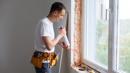 Eine Person mit Werkzeuggürtel steht in einem Raum neben einem Fenster. Um das Fenster sind die Wände und das Fensterbrett noch unverputzt. Die Person hält eine längliche Dose mit einem davon länglich abstehenden Stäbchen.