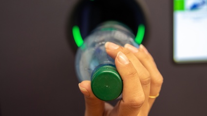 Nahaufnahme der rechten Hand einer Person, die eine Flasche horizontal in ein Loch hält. Im Loch sind innen am Rand drei grüne Lichter. Rechts vom Loch ist ein Bildschirm.