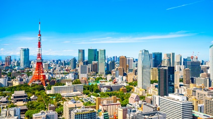 Stadtansicht von Tokio mit vielen Hochhäusern und dem bekannten rot-weißen Tokyo Tower unter blauem Himmel. 
