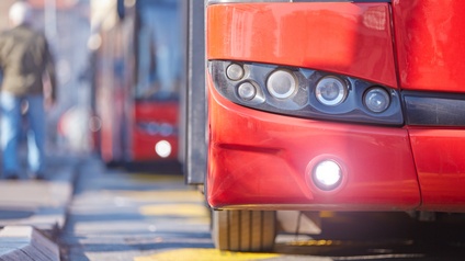 An einer Haltestelle halten hintereinander zwei rote Busse an einem Gehweg. Auf dem Gehweg steht eine Person