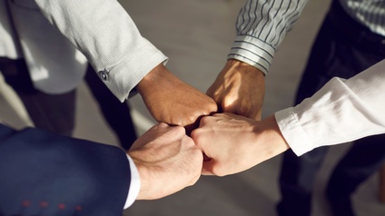 Detailaufnahme der linken Hände von 4 Personen, die zu vier Fäusten in der Bildmitte zusammentreffen, die Personen tragen Businesskleidung