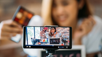 Im Vordergrund ist ein Smartphone horizontal in einer Halterung fixiert. Auf dem Bildschirm ist eine Person, die verschiedene Lidschatten in die Kamera hält. Vor der Person stehen weitere Schminkutensilien