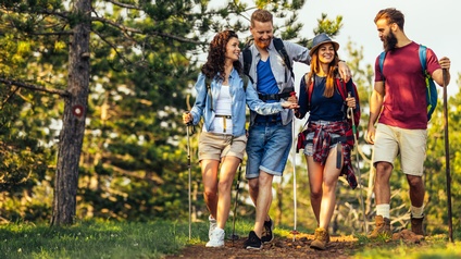 vier Personen, die nebeneinander im Wald gehen. Alle tragen Freizeitkleidung und Rucksäcke sowie Trekkingstöcke. Die Gruppe geht nebeneinander und blickt einander zu