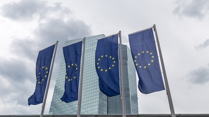 Vier europäische Flaggen sind an vier Masten befestigt und wehen im Wind. Hinter den Flaggen sieht man ein hohes, verglastes Gebäude