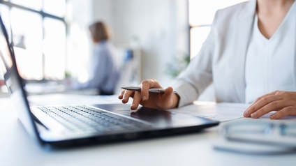 Schräge Aufnahme einer Person mit hellem Oberteil sowie hellem Sakko darüber, die an einem Tisch sitzt und vor sich einen aufgeklappten Laptop sowie einen Zettel hat. Im Hintergrund sitzt eine weitere Person an einem Tisch