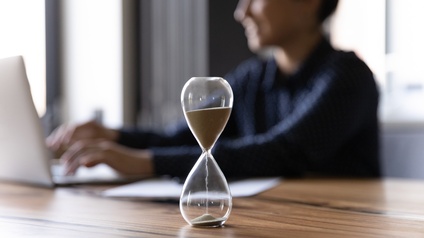 Eine Sanduhr steht auf einem Tisch. Die Sandkörner fließen durch die Öffnung. Im Hintergrund sitzt eine Person am Tisch. Sie blickt in einen aufgeklappten Laptop und hat ihre Hände auf der Tastatur liegen.