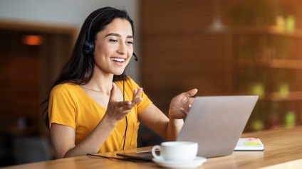 Person mit Headset und gelbem T-Shirt sitzt freudig vor einem Laptop und hält eine Konferenz, daneben steht eine Tasse sowie liegt ein Notizbuch auf dem Holztisch in einem Café