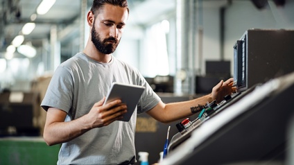 Eine Person steht vor einer CNC-Maschine und hält ein Tablet in der rechten Hand. Die Person schaut konzentriert auf das Tablet. Im Hintergrund ist eine Industriehalle zu sehen.
