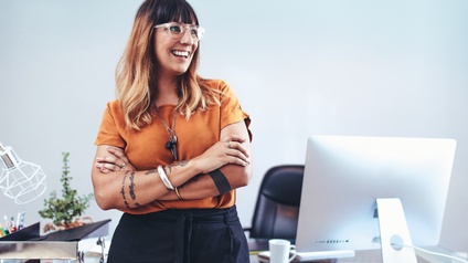 Frontale Aufnahme einer Person in einem orangen Oberteil, dunkler Hose, verschränkten Armen und Brille, die in den rechten Bildbereich blickt. Die Person lehnt sich an einem Schreibtisch hinter ihr an, auf dem ein Bildschirm und Büroutensilien sind.