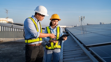 Man sieht zwei Personen, die auf einem Flachdach stehen, eine hält ein Tablet in der Hand. Man sieht Sonnenpanele und Hochhäuser im Hintergrund. 