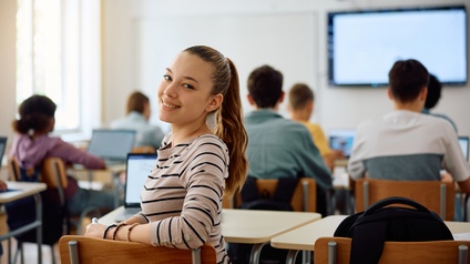 Jugendliche Person mit gestreiften Pullover sitzt an einem Tisch in einem Klassenzimmer mit Beamer-Projektion und blickt über ihre Schulter nach hinten in die Kamera