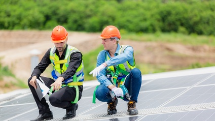 Zwei Personen in gelben Warnwesten mit orangen Schutzhelmen hocken auf Trittfläche auf Dach mit Solarpanelen und deuten auf bestimmte Stelle, im Hintergrund verschwommen belaubt Büsche