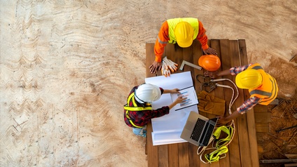 Baubesprechung mit drei Personen in Arbeits- und Schutzbekleidung stehen rund um einen Tisch mit Plänen, Laptops und weiteren Materialien von Topview-Perspektive in Baustellenumgebung